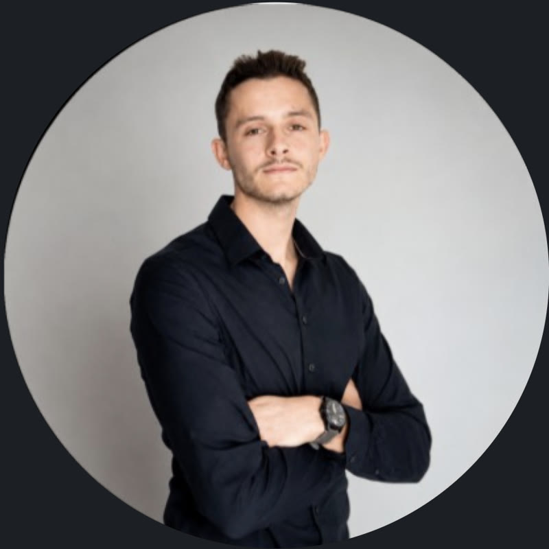 Portrait de Corto Benzoni, ergonome, chemise noire, bras croisés, sur fond gris.
