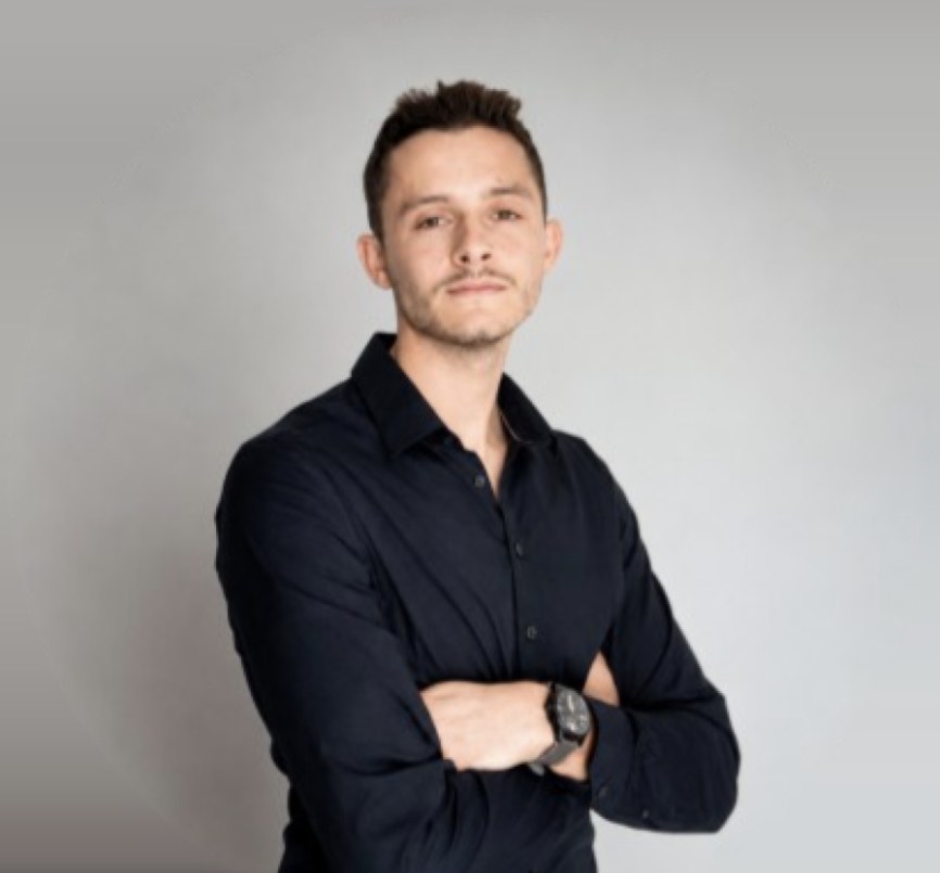 Portrait de Corto Benzoni, chemise noire, bras croisés, sur fond gris clair.